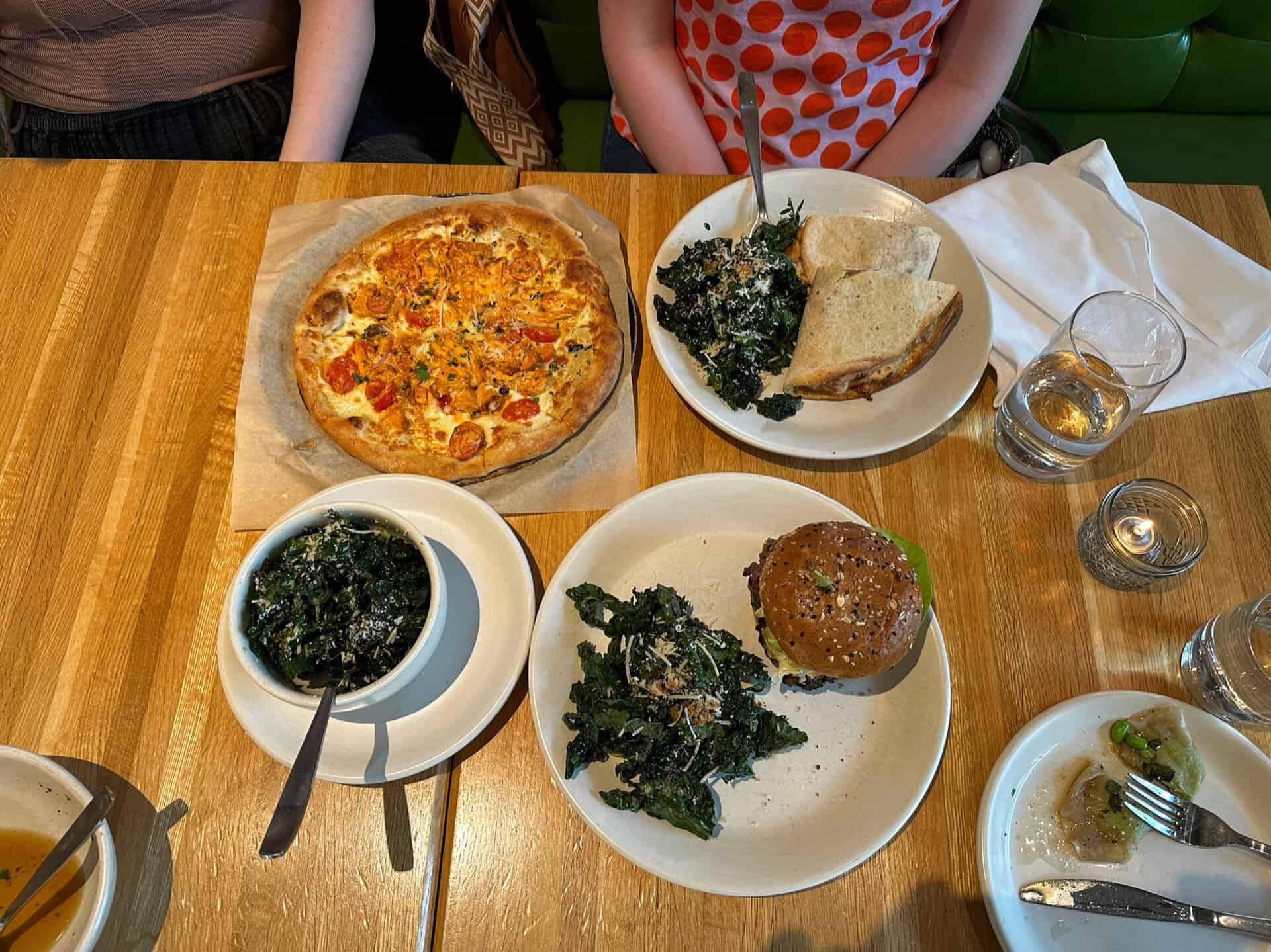 A Nashville-inspired table setting features a mouthwatering pizza, a burger topped with greens, a refreshing side bowl of greens, a plate of bread, and two glasses of water. Partially visible in the background are two seated people enjoying this delightful meal.
