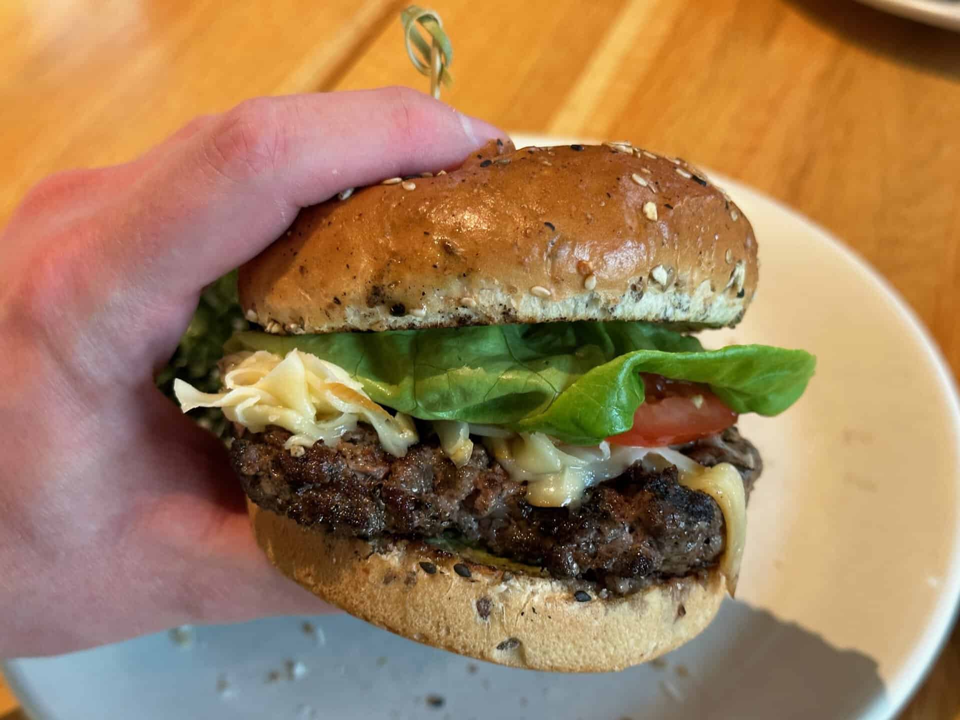 A hand holds a Nashville-inspired beef burger, brimming with lettuce, tomato, cheese, and nestled in a perfectly seeded bun on a white plate.