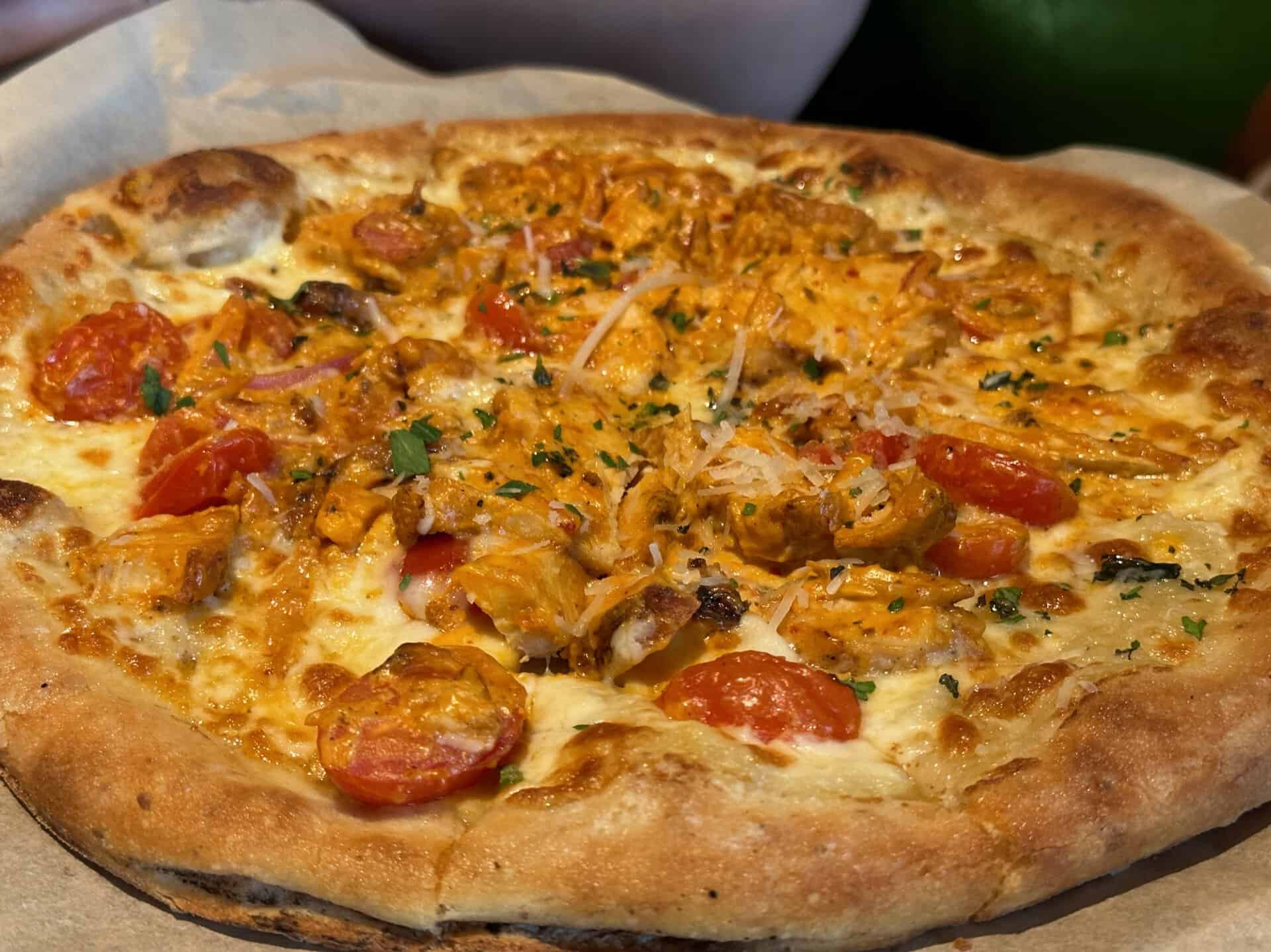 Close-up of a Nashville-style pizza topped with pieces of chicken, cherry tomatoes, herbs, and melted cheese, all resting on a parchment-lined tray.