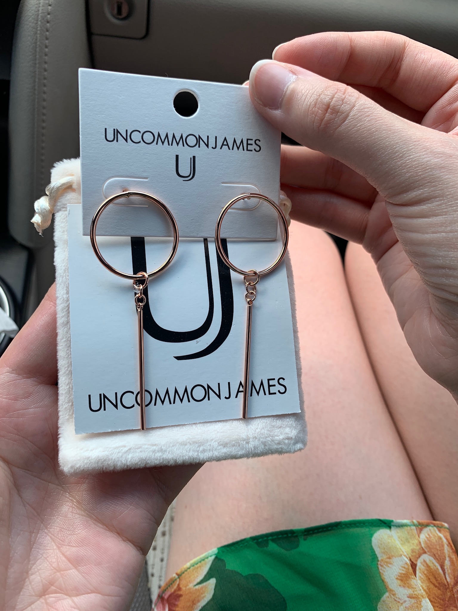 A hand holds a pair of gold hoop earrings with vertical bar pendants on an Uncommon James branded card, with a floral skirt visible in the background.