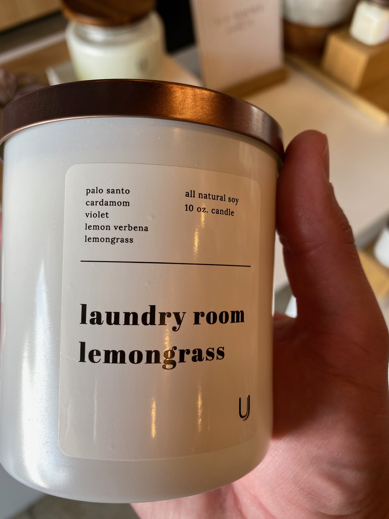 A hand holds a white soy candle labeled "laundry room lemongrass," listing scents including palo santo, cardamom, violet, lemon verbena, and lemongrass.