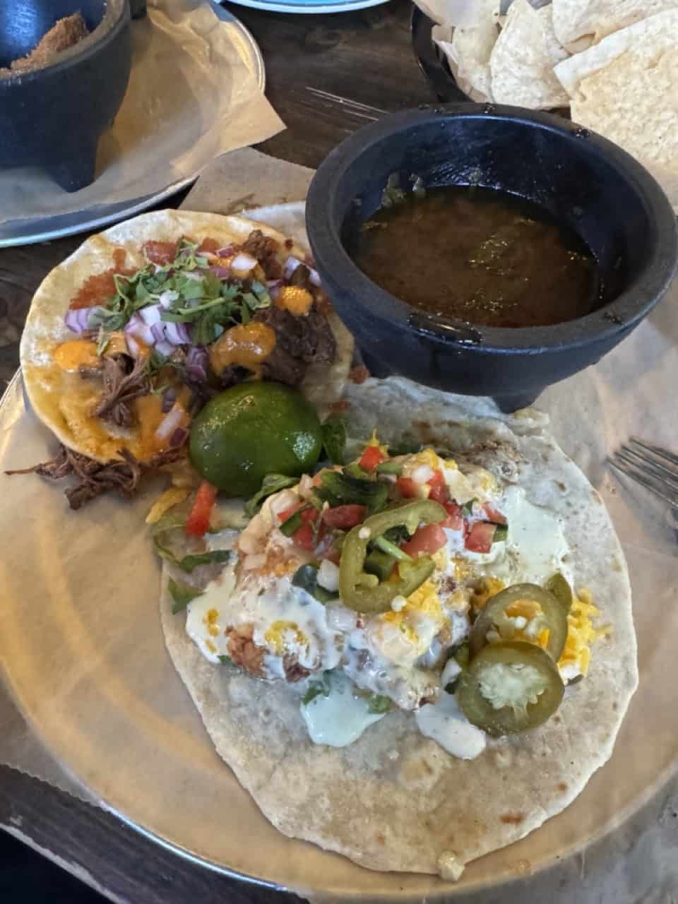 Two tacos on a plate, one with beef, onions, and cilantro, the other topped with jalapeños. A lime wedge and a bowl of green sauce accompany these Nashville-inspired delights. Chips linger in the background, completing this flavorful feast.