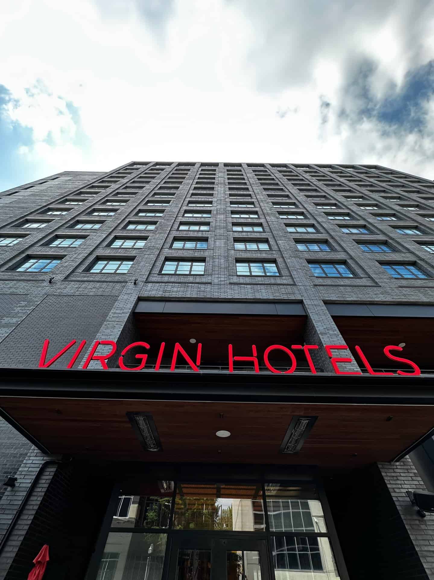 A tall brick building with a red "Virgin Hotels" sign above the entrance towers against the partly cloudy Nashville sky, captured from a low angle.