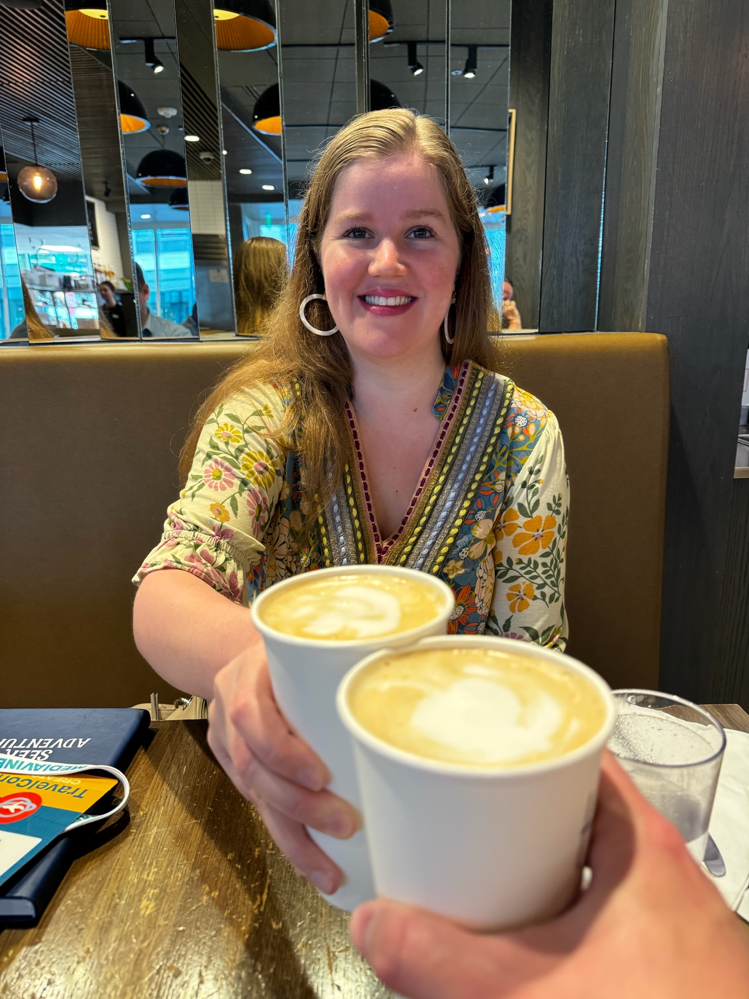 A woman smiles while sitting in a cafe booth, holding a cup of coffee and toasting with another person holding a similar cup.