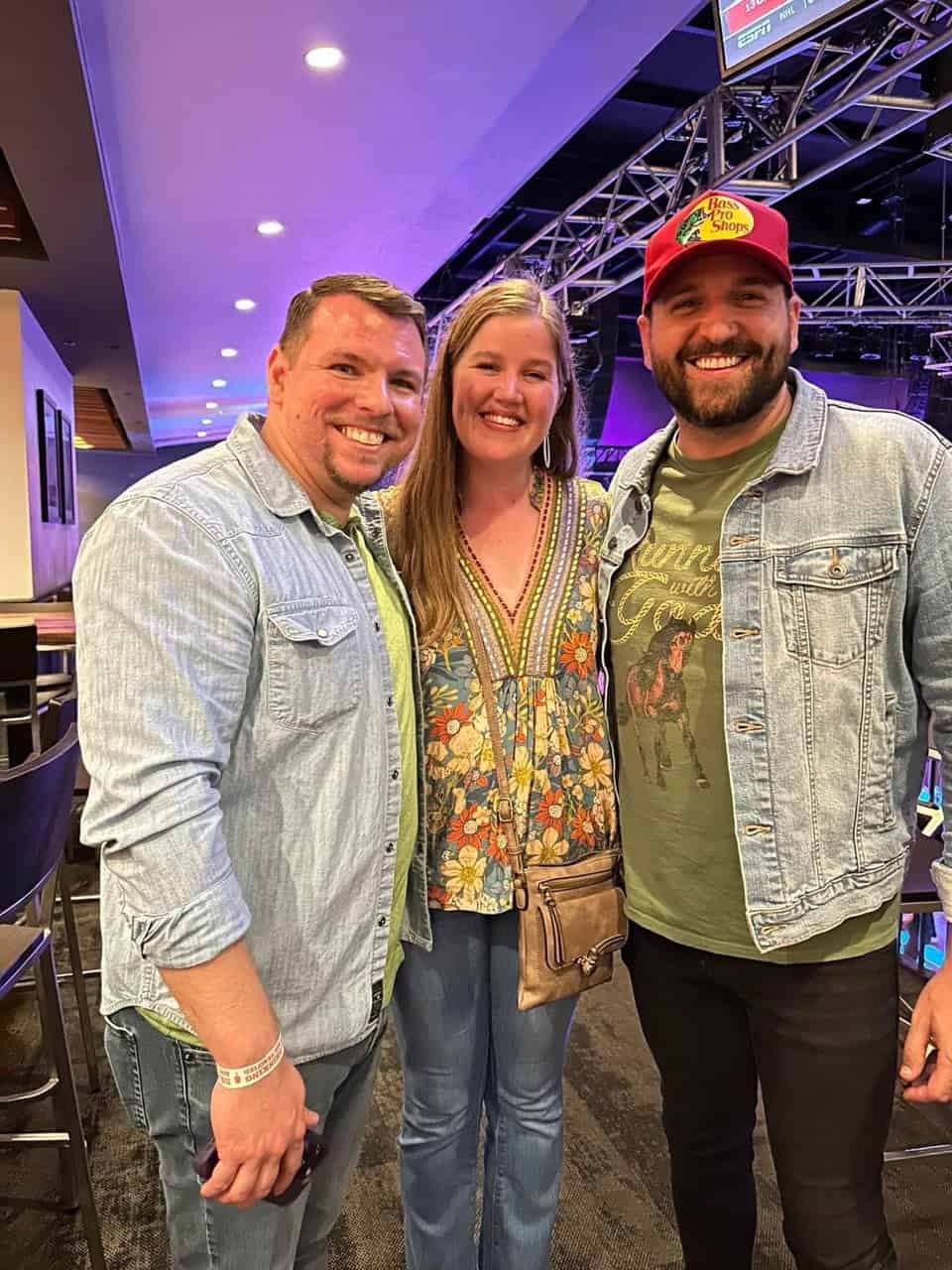Three people, two men and a woman, stand smiling in a casual Nashville venue. They are wearing jeans and casual shirts. The man on the right sports a red cap and denim jacket. Stage lighting is visible above.