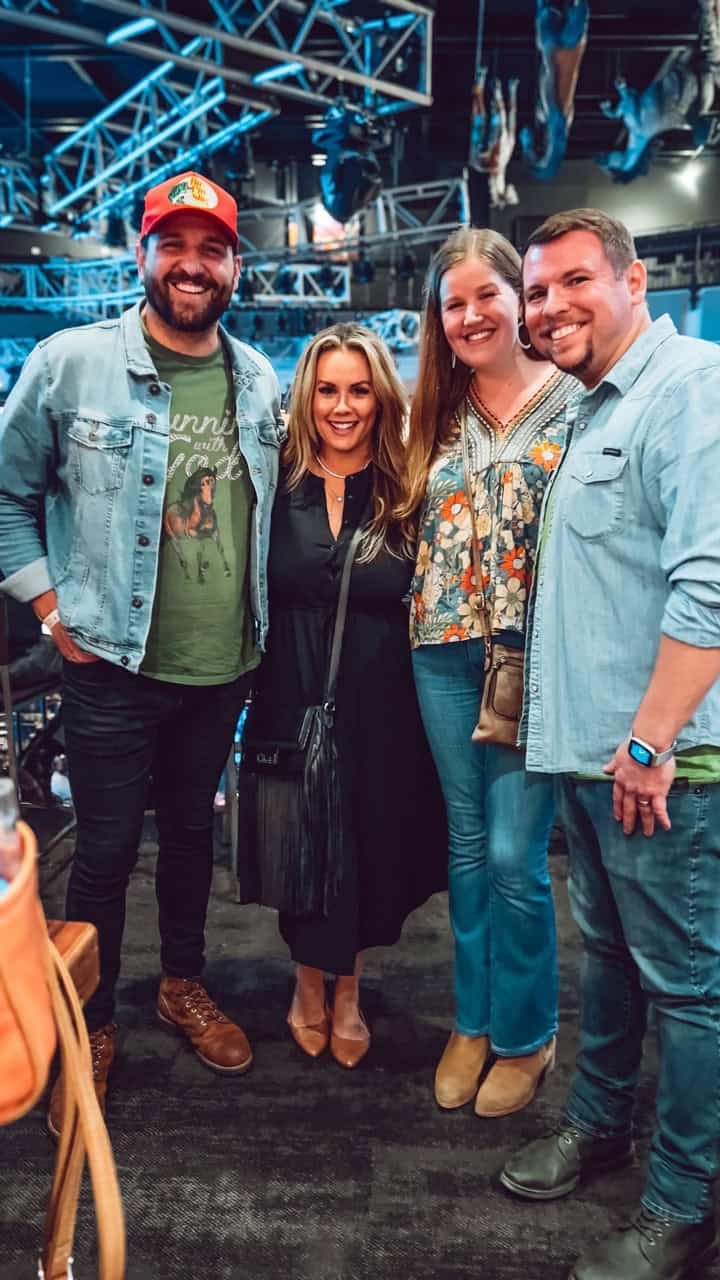 Four people stand together, smiling at the camera. The man on the left sports a red cap, while the woman in the middle dazzles in a black dress. With Nashville vibes in the air, stage lights glow warmly above their cheerful indoor gathering.