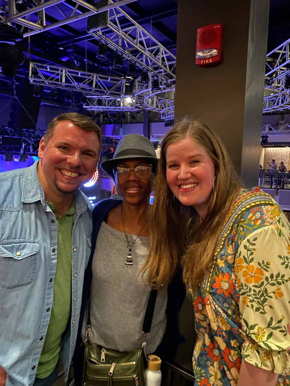 Three people smiling indoors at a Nashville venue with stage lighting in the background. The person in the middle wears a hat and glasses.