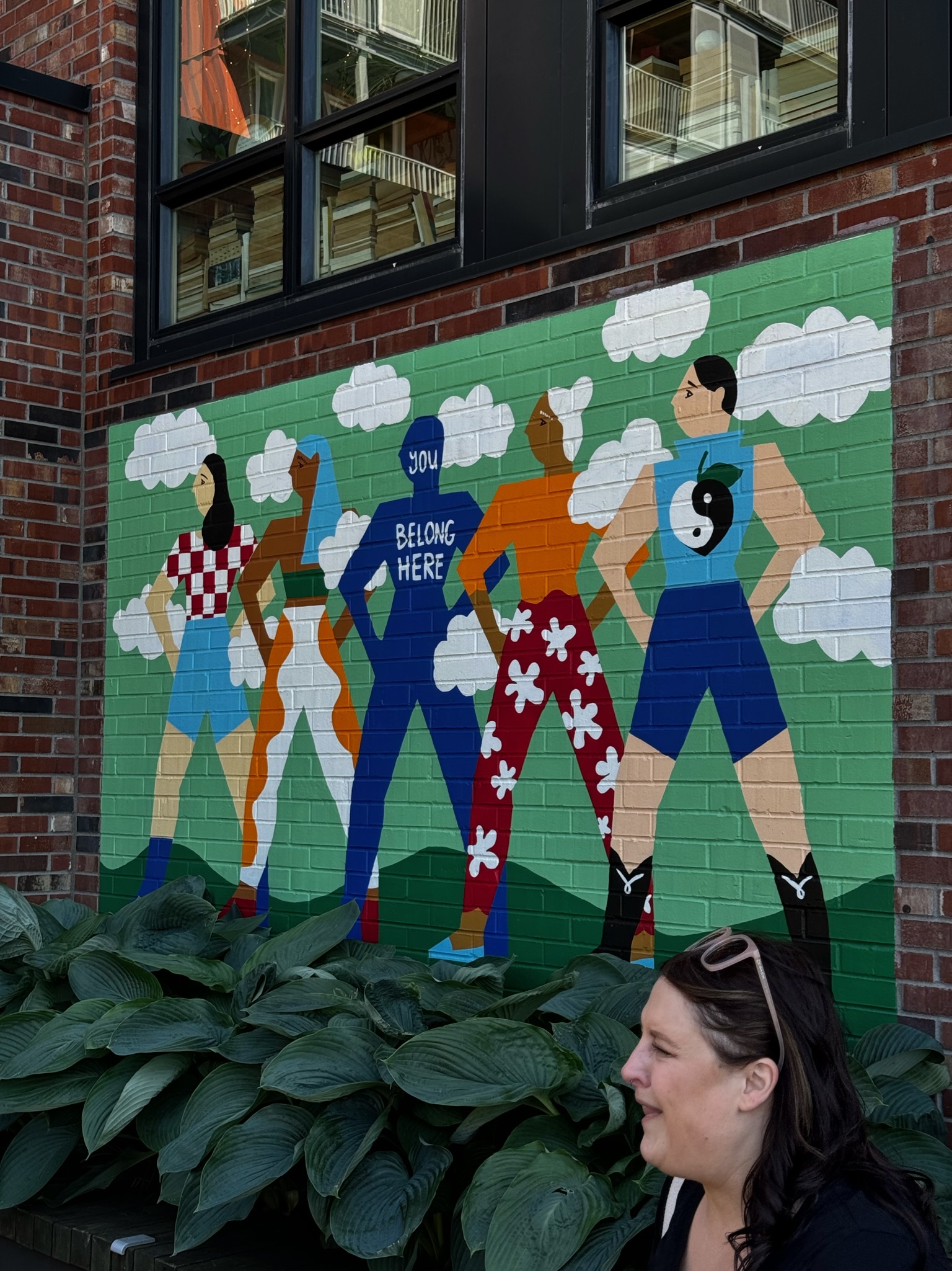 A mural on a brick wall shows five diverse figures standing with hands on hips; one has "You Belong Here" on their shirt. Green plants and a woman are in the foreground.